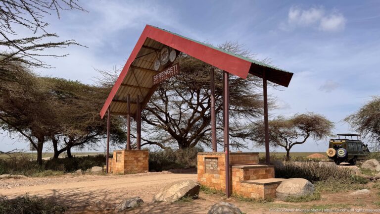 Serengeti National Park entrance with red roof and tall posts beside a dirt road, with a safari vehicle parked at right.