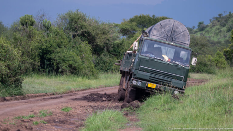 Green safari truck tilted into a deep muddy rut on a narrow track bordered by dense vegetation.