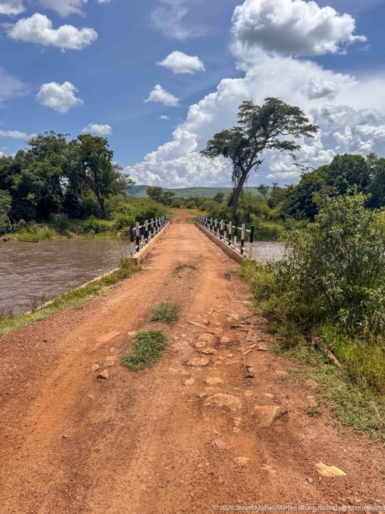 Red dirt road crossing a low bridge toward green woodland and a broad flat-topped tree under bright clouds.