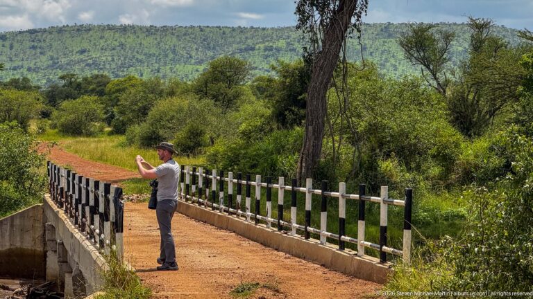 Person standing on a narrow dirt bridge holding up a camera or phone, surrounded by dense green vegetation.