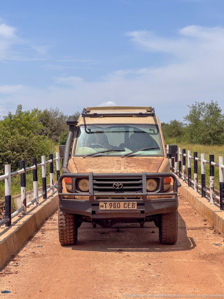 Dusty Toyota Land Cruiser safari vehicle facing the camera on a narrow bridge with black-and-white rail posts.