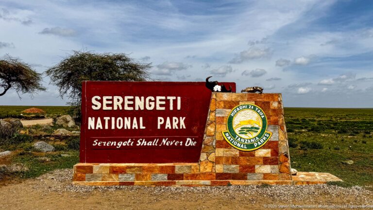 Serengeti National Park red-and-stone entrance marker standing alone against open plains and a blue, cloud-dotted sky.