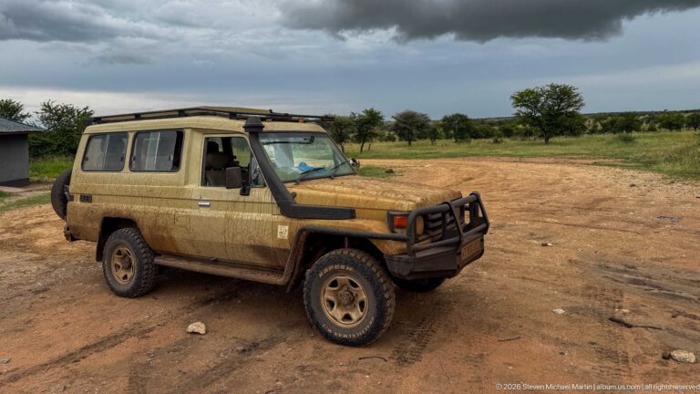 Mud-covered four-wheel-drive safari vehicle parked on bare ground beneath dark storm clouds, with grassland and trees beyond.