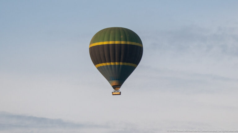 Distant dark hot-air balloon with yellow bands floating above a hazy horizon under a pale sky.