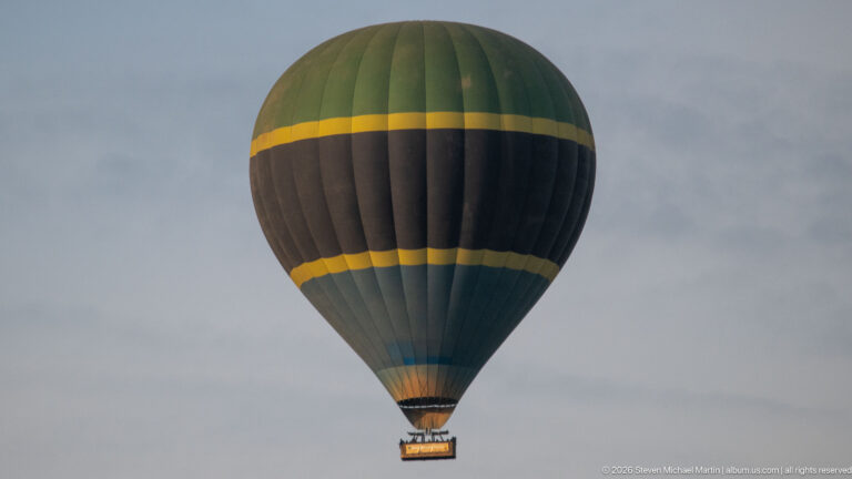 Dark hot-air balloon with two yellow bands floating against a gray-blue sky, passenger basket suspended below.