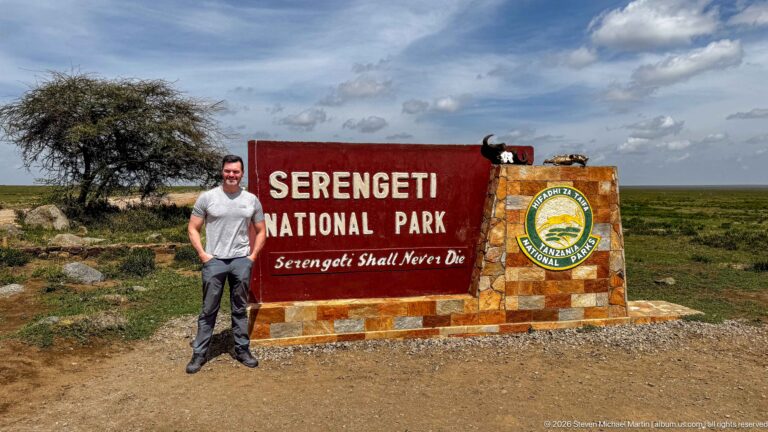Single visitor beside the Serengeti National Park stone entrance marker with red sign and TANAPA emblem.