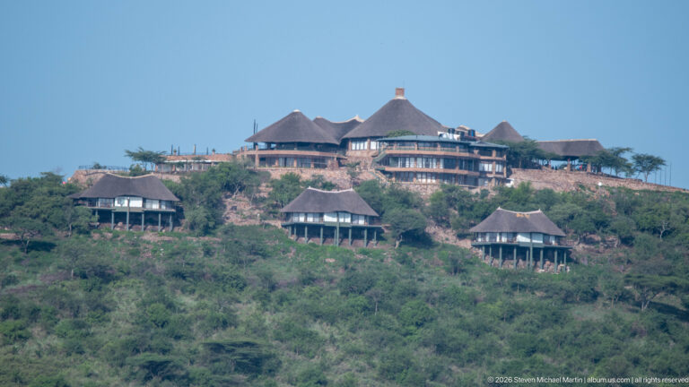 Large safari lodge and smaller pitched-roof buildings spread across the crest and upper slope of a green hill.