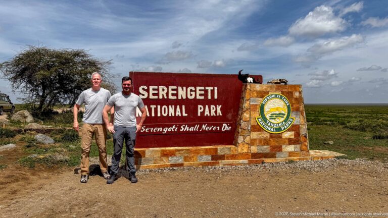 Two visitors standing beside the red-and-stone Serengeti National Park entrance marker on an open plain.