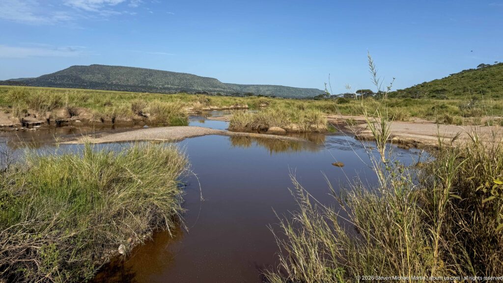 Shallow brown-blue pool edged by marsh grass and sandy banks, with low green hills under a clear sky.