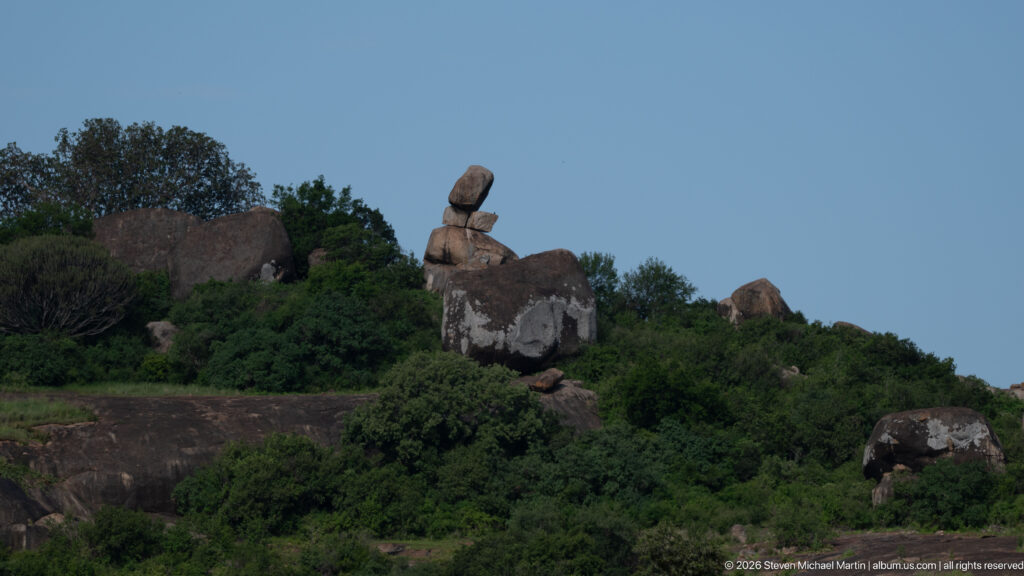 Naturally balanced granite boulders stacked on a shrub-covered Moru Kopjes slope beneath blue sky.