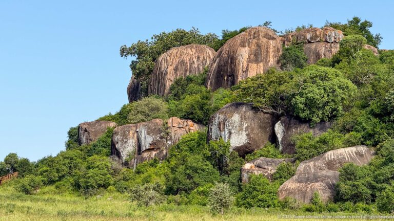 Massive rounded granite boulders with dark weathering streaks and dense shrubs above green grassland.