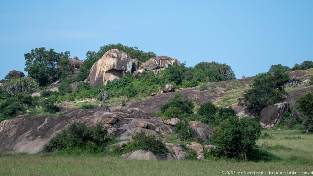 Large rounded granite boulders and smooth rock slabs rising among shrubs above Serengeti grassland.
