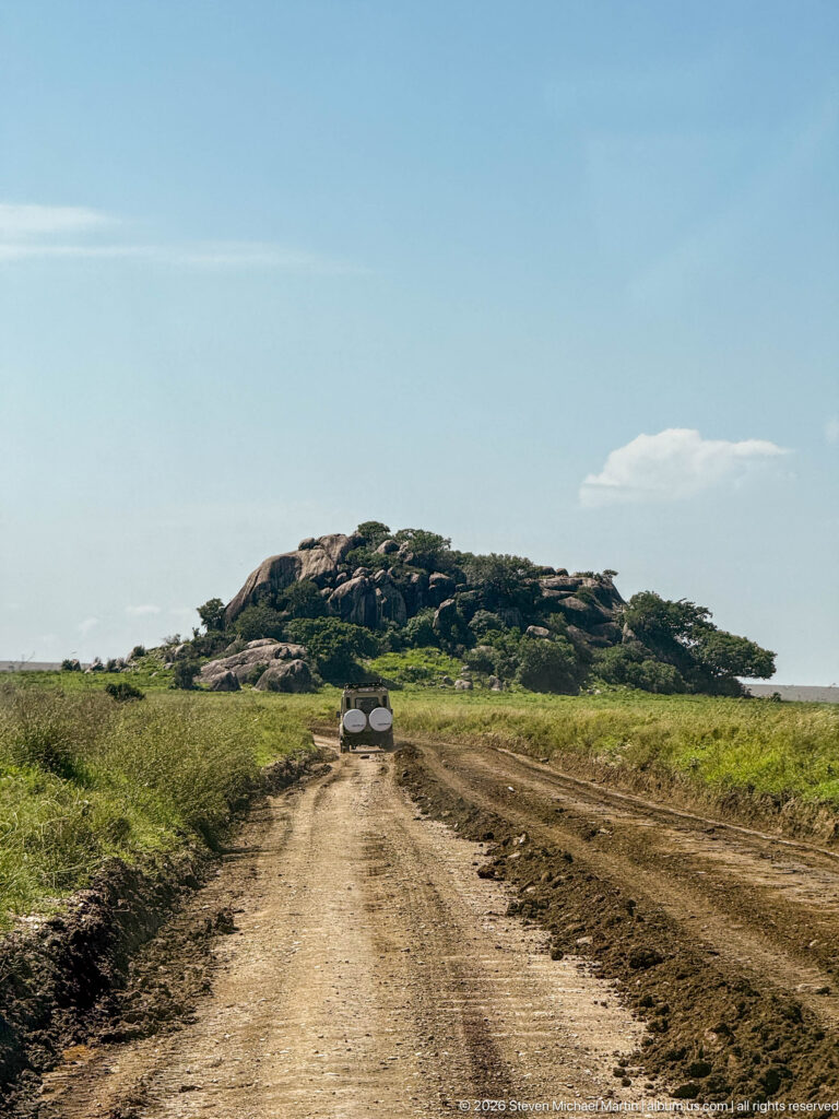 Dirt safari road crossing green grassland toward a low granite kopje with a vehicle ahead.