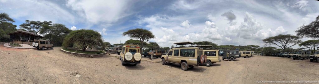 Multiple safari four-wheel-drives parked on a gravel lot beside low buildings and scattered trees under large clouds.
