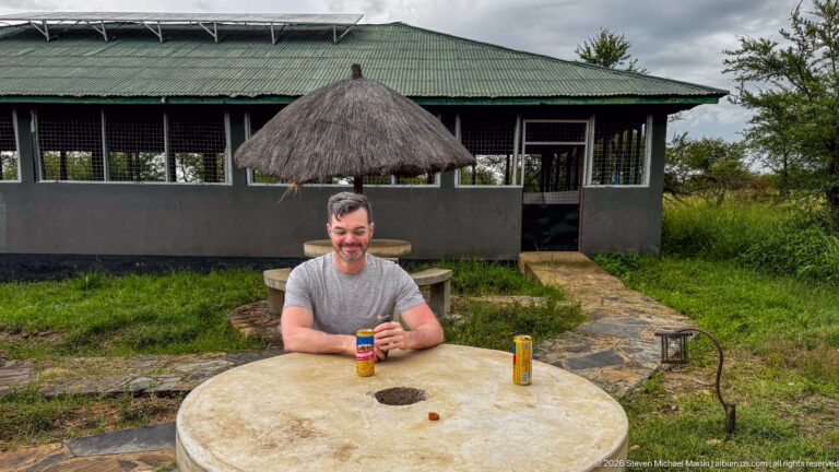 Traveler seated at a round outdoor table with drink cans beside a lodge building and thatched shade.