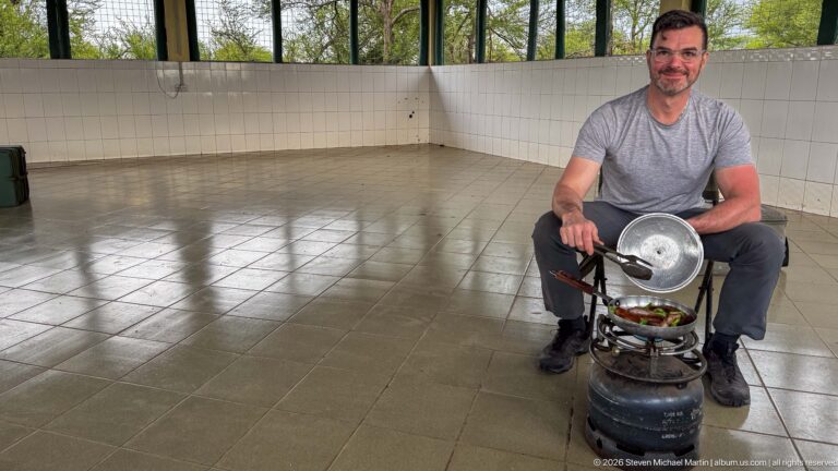 Traveler seated in a tiled shelter, tending a pan on a compact stove between his feet.