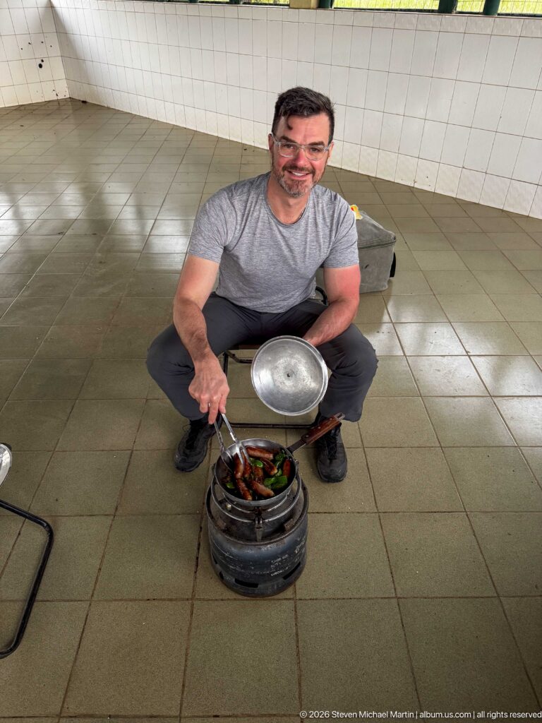 Traveler crouching over a small charcoal stove with a pan of food in a tiled open-sided shelter.