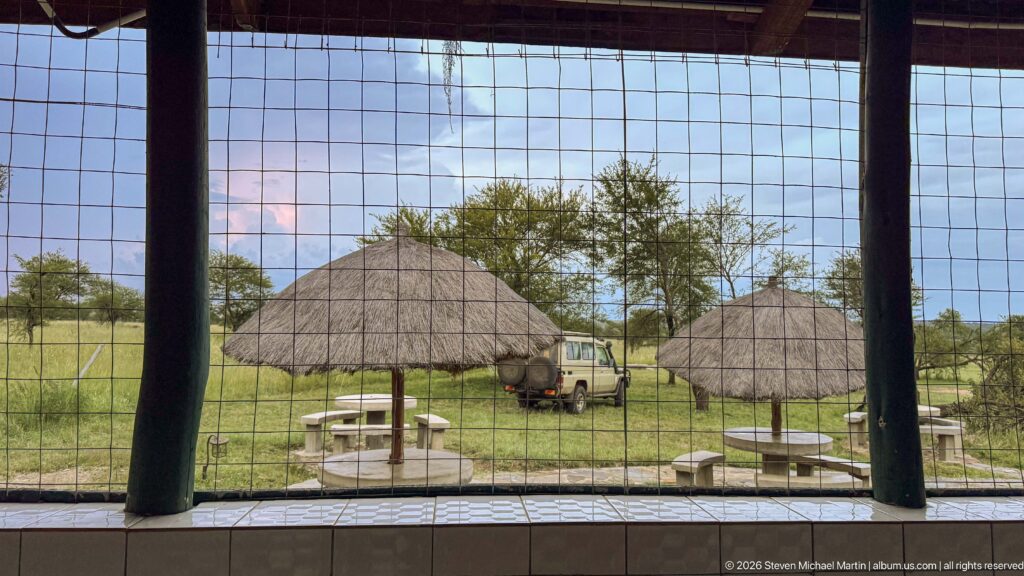 View through square wire mesh toward thatched picnic shelters, stone tables, grass, trees, and a parked safari vehicle.