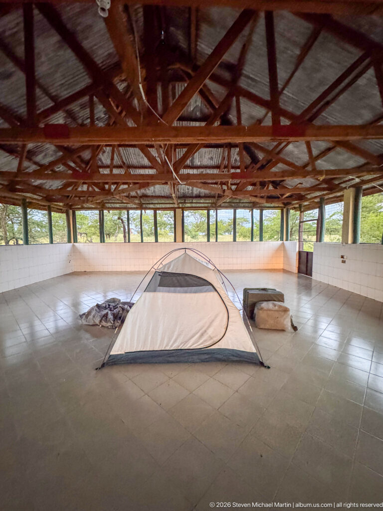 Small dome tent and bedding on a tiled floor inside a large open-sided shelter with exposed timber roof trusses.