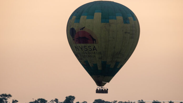 Large Nyssa Balloon Safaris hot-air balloon with passenger basket floating above the Serengeti in warm light.
