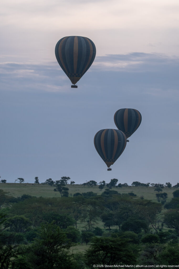 Three hot-air balloons floating above a green, tree-dotted Serengeti savanna beneath a pale morning sky.