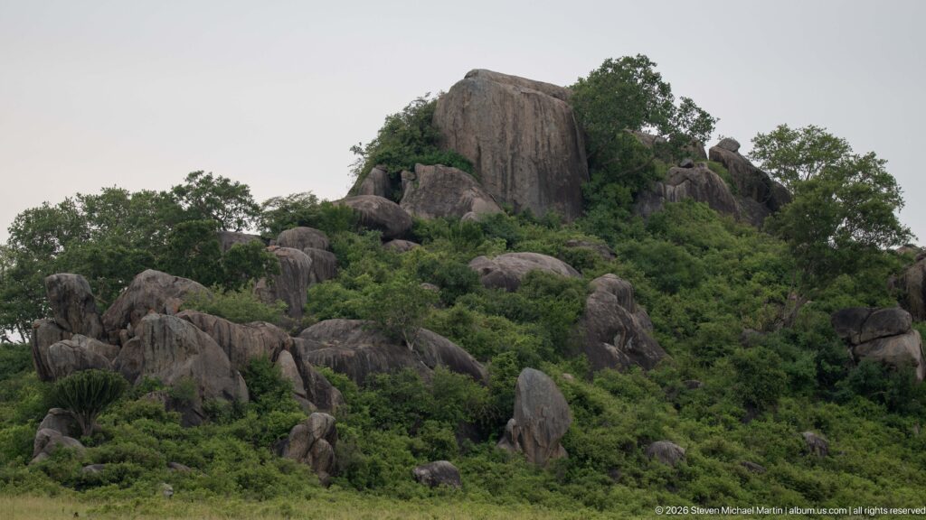 Large upright rock tower surrounded by rounded boulders and dense shrubs on a Simba Kopjes slope.