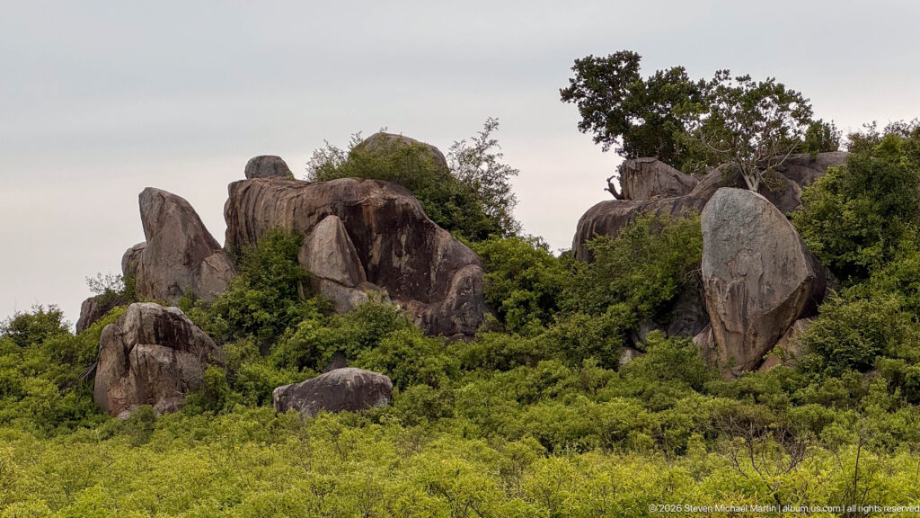 Clustered weathered boulders and steep rock slabs rising above dense green shrubs with trees rooted among them.