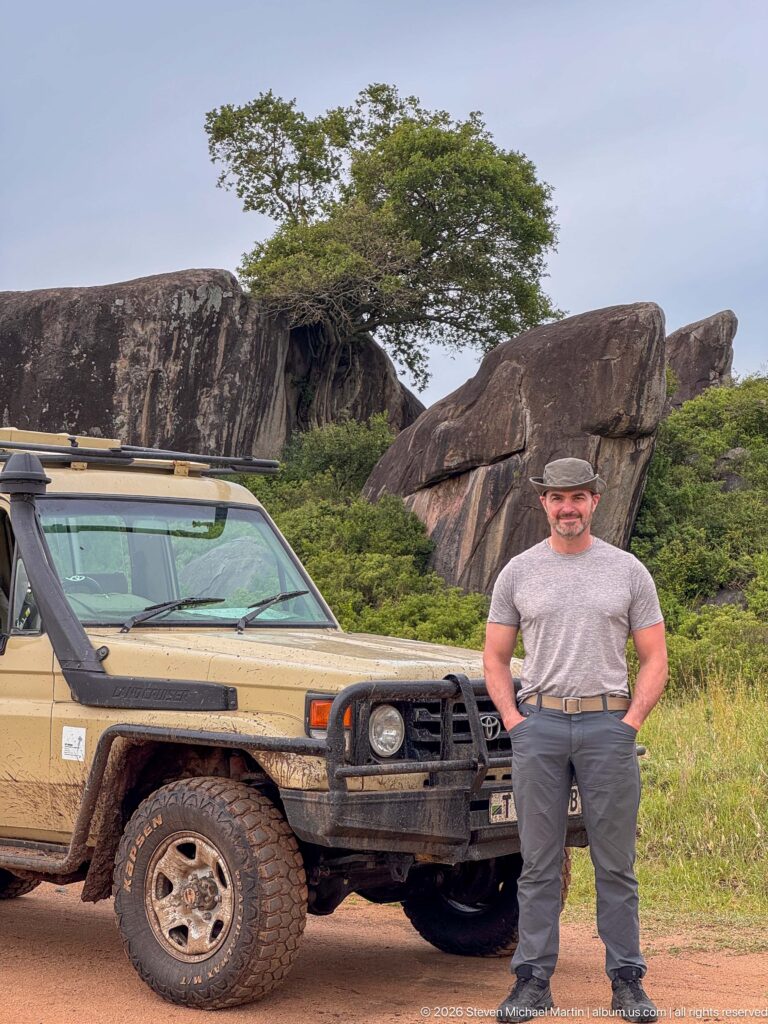 Traveler standing before a safari vehicle with massive rounded and fractured kopje boulders behind.