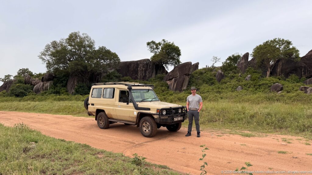 Person standing beside a tan safari vehicle on a dirt road, with large rounded rock outcrops and trees behind.