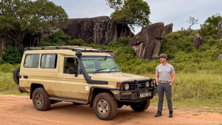Traveler beside a safari vehicle on a dirt track below tall weathered kopje rock slabs.
