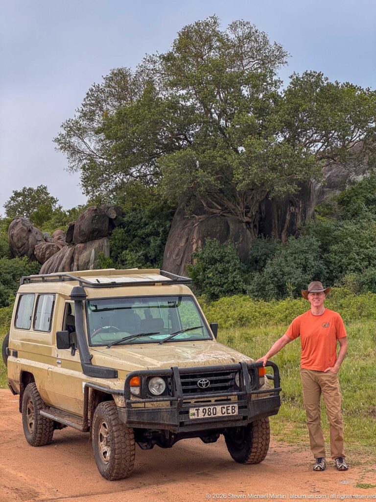 Traveler standing beside a safari vehicle in front of large rounded boulders and green vegetation at Simba Kopjes.