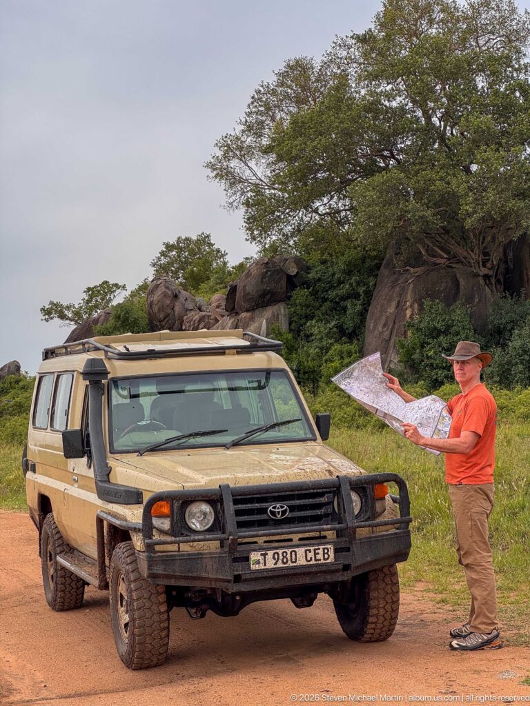 Traveler reading a large paper map beside a safari vehicle with rounded Simba Kopjes rocks behind.