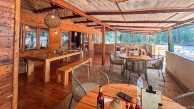 Wooden open-air café with tables, woven chairs, benches, and forest visible beyond the railing.