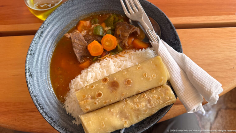 Bowl of meat and vegetable stew with white rice and folded chapati on a wooden table.