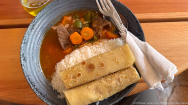 Bowl of meat and vegetable stew with white rice and folded chapati on a wooden table.
