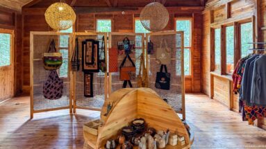 Wooden lodge gift shop displaying bags, textiles, clothing, woven lamps, and small souvenirs.