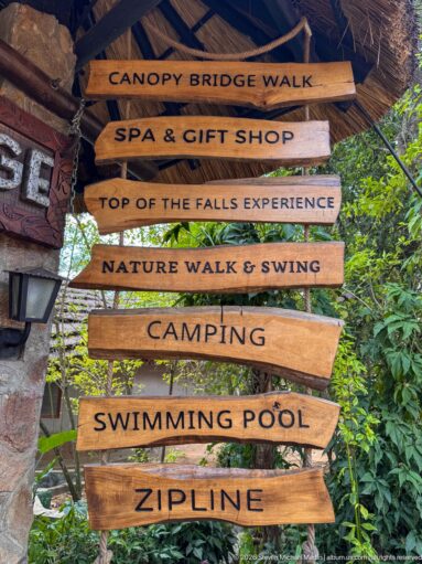 Stacked wooden directional signs listing canopy bridge walk, spa, nature walk, camping, swimming pool, and zip line.