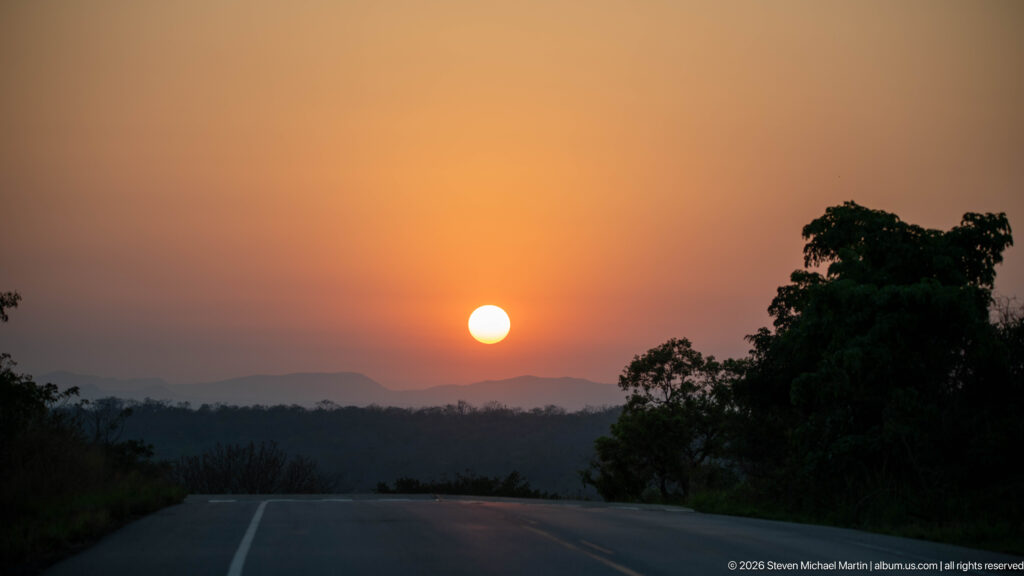 Low sun over a paved road, dark forest canopy, and hazy distant ridge near Budongo Forest.