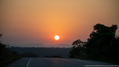 Low sun over a paved road, dark forest canopy, and hazy distant ridge near Budongo Forest.