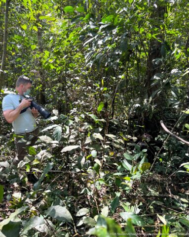Photographer holding a long-lens camera while standing in dense green understory and tangled forest stems.