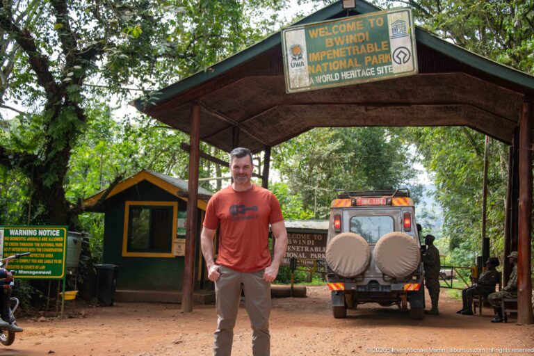 Full-length man beneath the Bwindi entrance gateway with checkpoint booth, warning sign and safari vehicle behind him.
