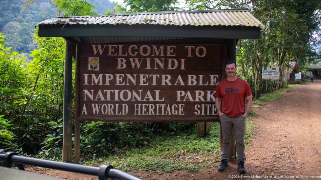 Man standing beside a wooden Welcome to Bwindi Impenetrable National Park, A World Heritage Site sign on a forest road.