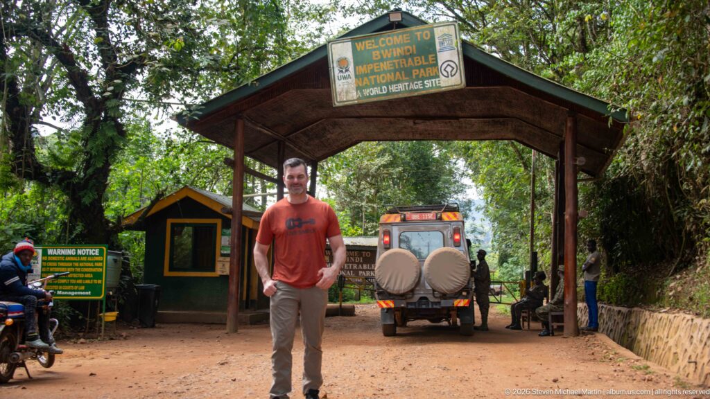 Man centered beneath the Bwindi park entrance canopy with a safari vehicle directly behind him on the dirt road.