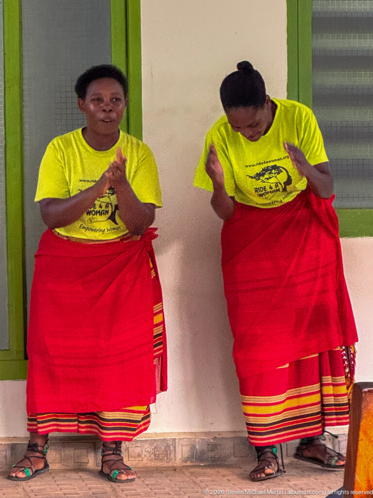 Two women in matching yellow shirts and red striped wrap skirts clap and dance side by side on a veranda.