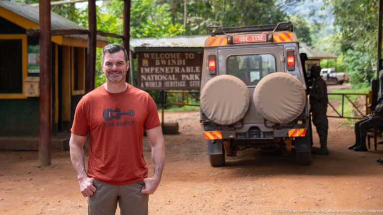 Smiling man near the Bwindi entrance with a safari vehicle and wooden national park welcome sign behind him.