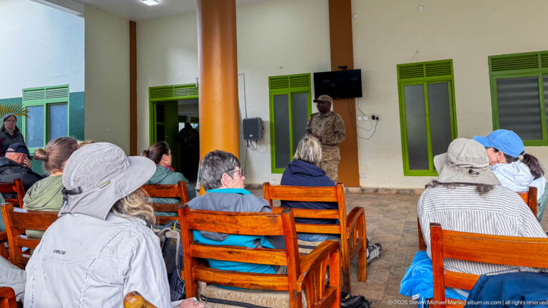 Seated visitors listen to a standing guide during a briefing on a veranda with green-trimmed doors and windows.