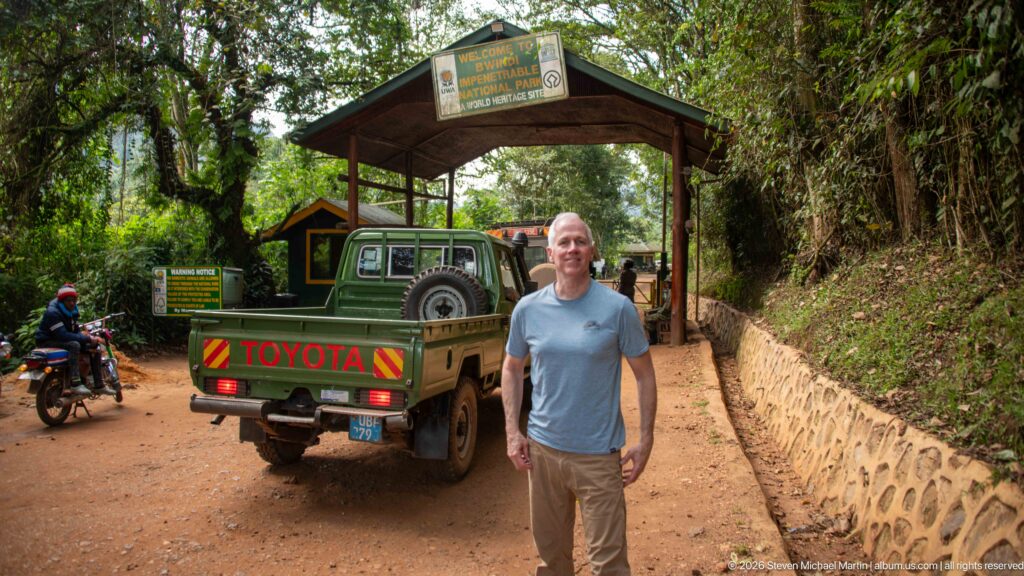 Man standing in front of a safari vehicle beneath the Bwindi Impenetrable National Park entrance gateway.