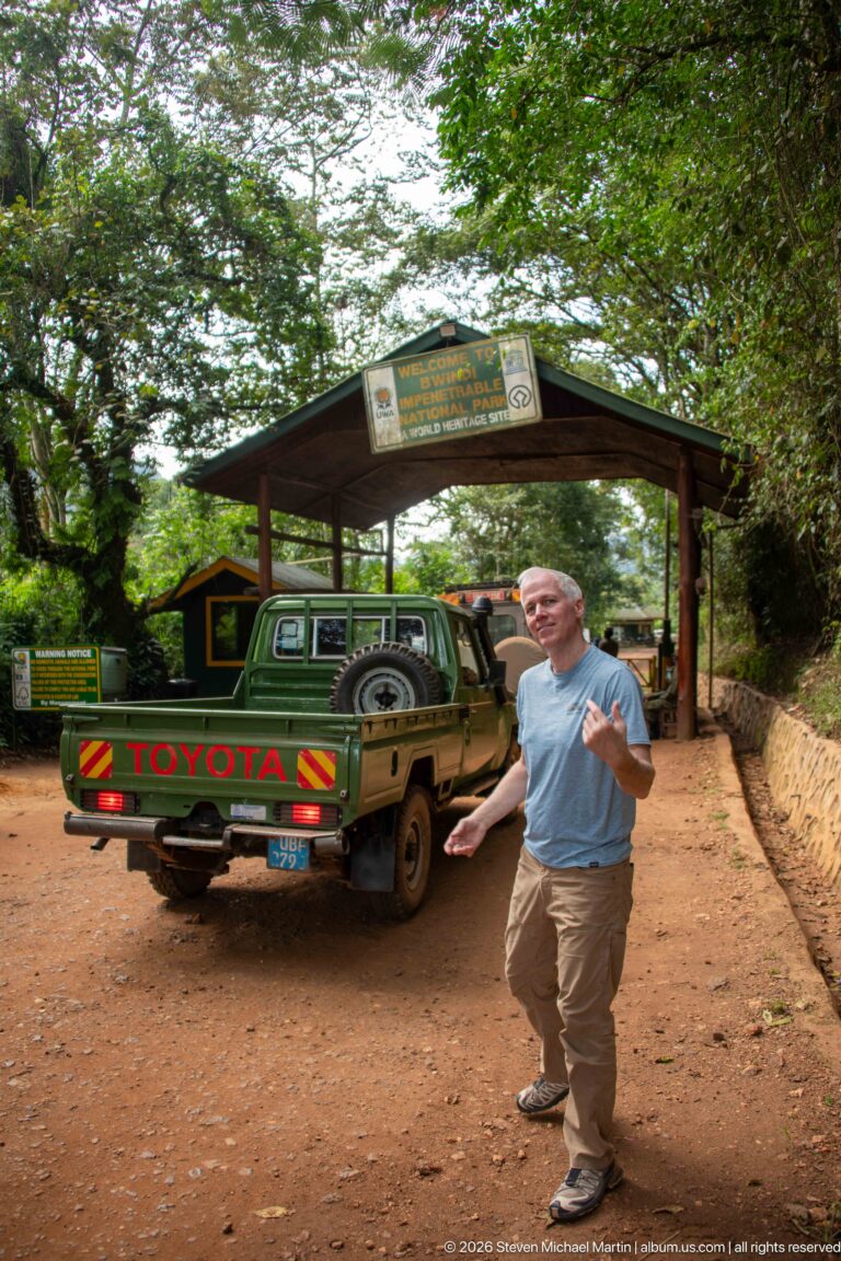 Man gesturing beside a safari vehicle under the covered entrance gateway to Bwindi Impenetrable National Park.