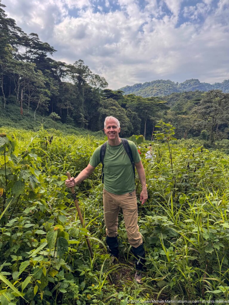 Trekker with walking stick standing in tall vegetation before layered, forested hills beneath a partly cloudy sky.