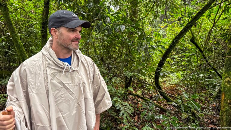 Man in a pale rain poncho and cap stands amid dense vines, branches, ferns, and green rainforest foliage.
